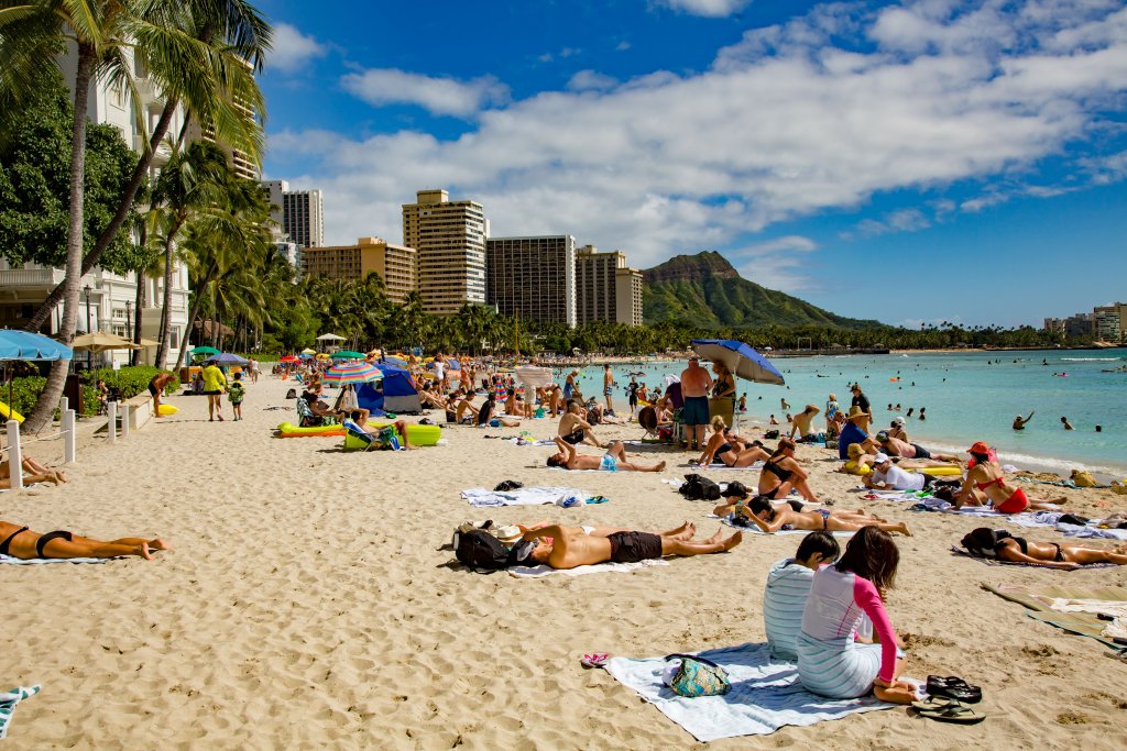 Crowded, sunny beach with high-rise hotels on the left and bright blue ocean on the right.